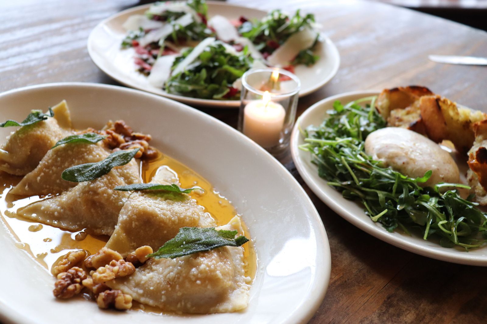 Plates of fresh pasta and burrata on a wooden table lit by a candle