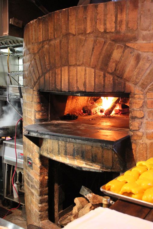 A wood-fired brick oven with an open flame inside, the mouth of the oven blackened from years of use